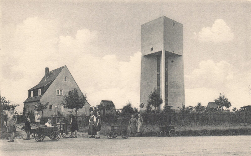 Der Wasserturm wirkt wie ein Fremdkörper in der ländlichen Bebauung mit Frauen und Kindern mit Holzwagen im Vordergrund.