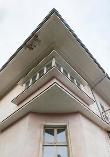 Foto an einer Gebäudeecke hinauf bis unter das Dach. Im unteren Bereich Ausschnitt der Bauecke, darüber springen Kragplatten des Fensters und vor allem das Dach weit hervor.