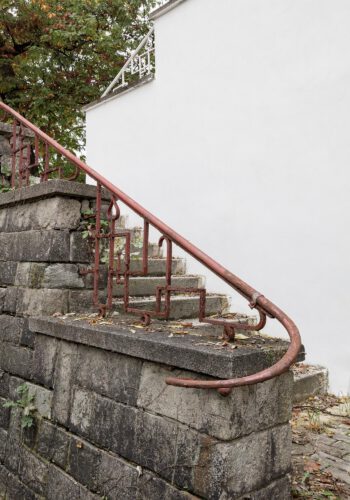 Ein rostbraunes Metallgeländer folgt den Treppenstufen auf einer Brüstungsmauer mit geometrischen Mustern und schwingt mit seinem Handlauf nach links. Dahinter die weiß geputzte Hauswand.