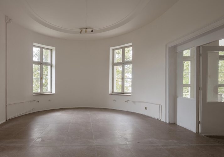 Foto eines leeren Raumes mit elipsenförmigen Grundriss und zwei Fenstern. Rechts führt eine große Tür in einen weiteren Raum.