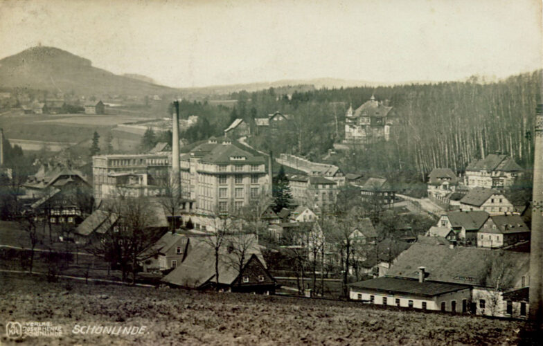 Der Ortsausschnitt liegt in einer leichten Senke. Etwa in der Mitte ein größerer Fabrikbau, rechts die alte Villa Palme, links im Hintergrund ein bewaldeter Berg hinter Feldern.