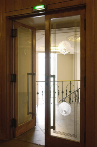 Eine zweiflügelige hohe Glastür mit kreisförmigen Ätzungen gibt den Blick auf das helle Treppenhaus mit vergoldetem Stuck und das leichte Metallgeländer fei.