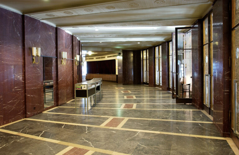 Der Blick in das Foyer zeigt einen viertelkreisfömigen Ausschnitt des Umgangs. Die violettbraunen Marmorwandverkleidungen, der Deckenstuck, der mehrfarbige Steinboden und die großen Außentüren sind als Kreissegmente miteinander verbunden.