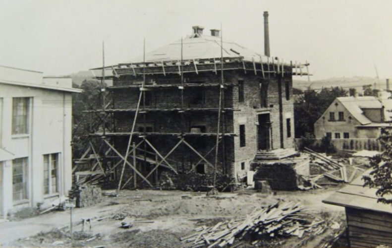 Foto von erhöhtem Standpunkt auf den eingerüsteten Rohbau der Villa, dessen Dach bereits geschlossen ist. Links neben der Villa eine Fabrikhalle, rechts dahinter ein älteres Gebäude.