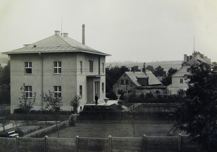 SW-Foto über einen Betonzaun und einen Garten mit Rasen und dünnen Bäumen hinweg auf das Haus, auf dessen Eingangsterrasse ein Mann steht. Hinter der Villa zwei ältere Häuser, dann ansteigende Landschaft. Schwarzweiß-Foto.