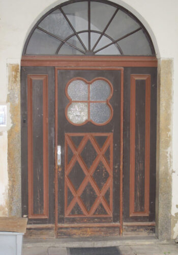Große Bogentür in Holz mit Rautenmuster und kleeblattartigem Fenster im Mittelteil sowie Fenster im Bogenabschluss.