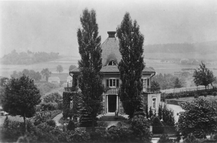 Das Foto von erhöhter Position zeigt das Wohnhaus hinter zwei mächtigen Pappeln und links einen berankten Erker.