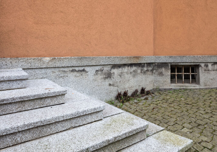 Foto von den von links schräg ins Bild ragenden Treppenstufen zum Eingang. Rechts dahinter der erhaltene Terrazzosockel unterhalb der orangefarbenen Putzwand.