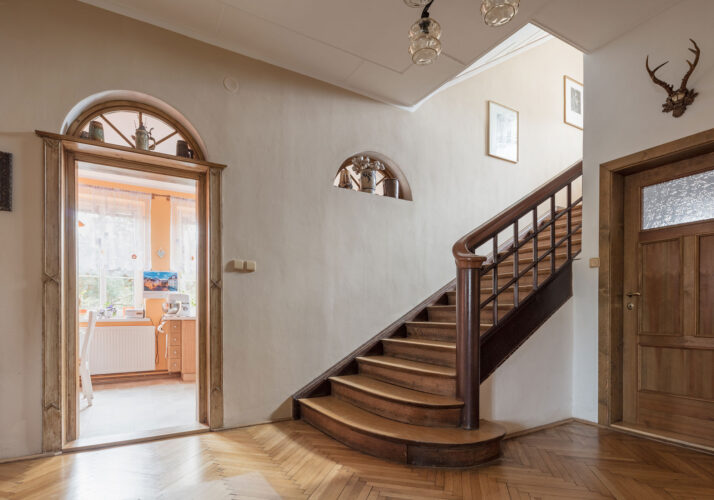 Foto von der Eingangshalle auf eine geöffnete Tür, die in einen Wohnraum führt, und eine Treppe, die nach oben führt. Parkettboden, hölzerne Treppe, Holztür mit halbrunden Oberlichtern.