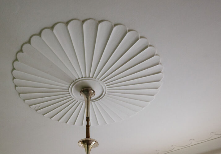 Fotografie štukovitého obrazce na stropu připomínajícího ventilátor z prostředka visí lustr.